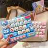 A hand holds a blue cartoon animal keyboard keycap fidget toy with various animal shapes and a pink keychain in a beige bag.