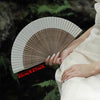 A close-up of a white bamboo folding fan with decorative accents held by a person in a natural, dark green outdoor setting.