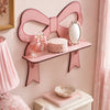 A pink wooden wall shelf shaped like a bow holds a mirror and a glass jar on a light pink wall.