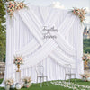 A white fabric backdrop with an 'X' design draped in soft folds and decorated with floral arrangements.