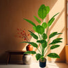 A large artificial tree with lush green leaves stands in a pot on a wooden table next to a window.