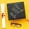 A black graduation card with silver lettering for Class of 2024 and Molly, next to a white rolled paper and black glasses.