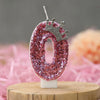 A pink sequin number zero candle with a silver crown topper displayed on a wooden surface.