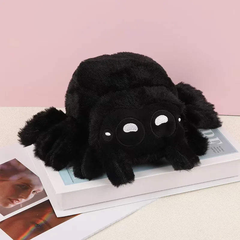 A black plush toy of a spider with large eyes is displayed on a white box next to some papers.