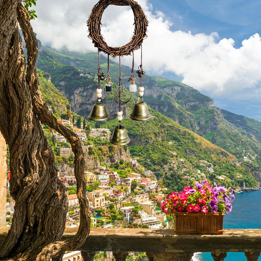 A rustic wreath of branches hangs from a rope, featuring several brass bells and decorative elements against a scenic landsca