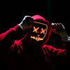 A person wearing a red hooded garment with a glowing orange LED mask featuring crossed eyes and a grinning mouth.