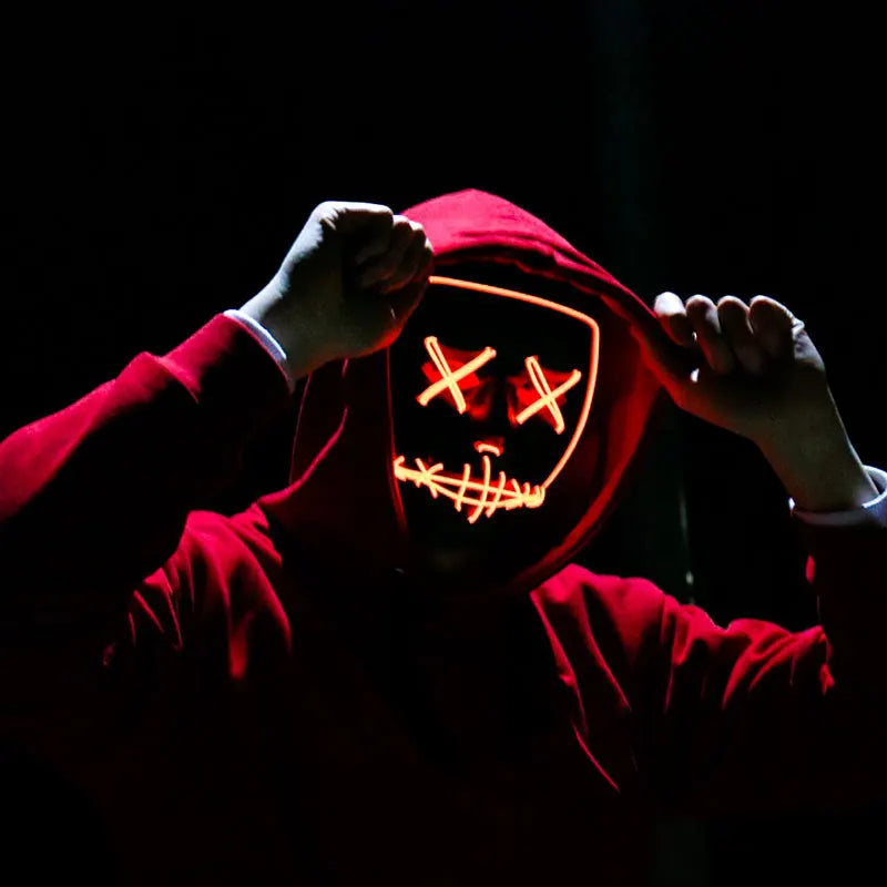 A person wearing a red hooded garment with a glowing orange LED mask featuring crossed eyes and a grinning mouth.