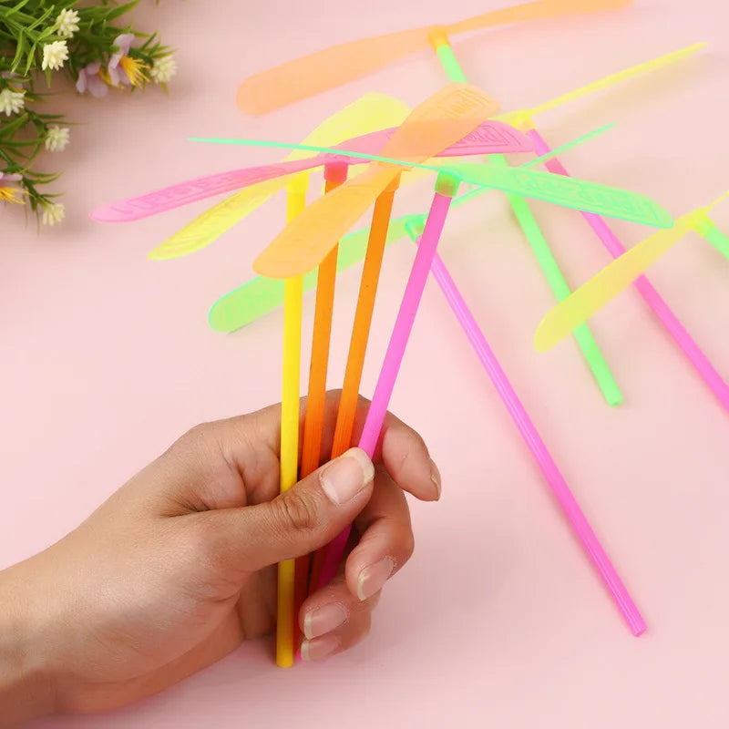 A hand holds a set of colorful bamboo dragonfly flying propeller toys against a pink background.