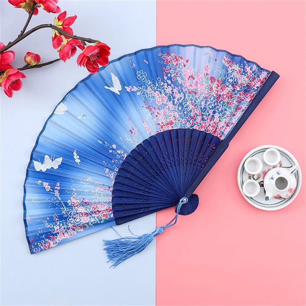 A blue and pink floral silk folding hand fan displayed on a pink background next to small white teacups.