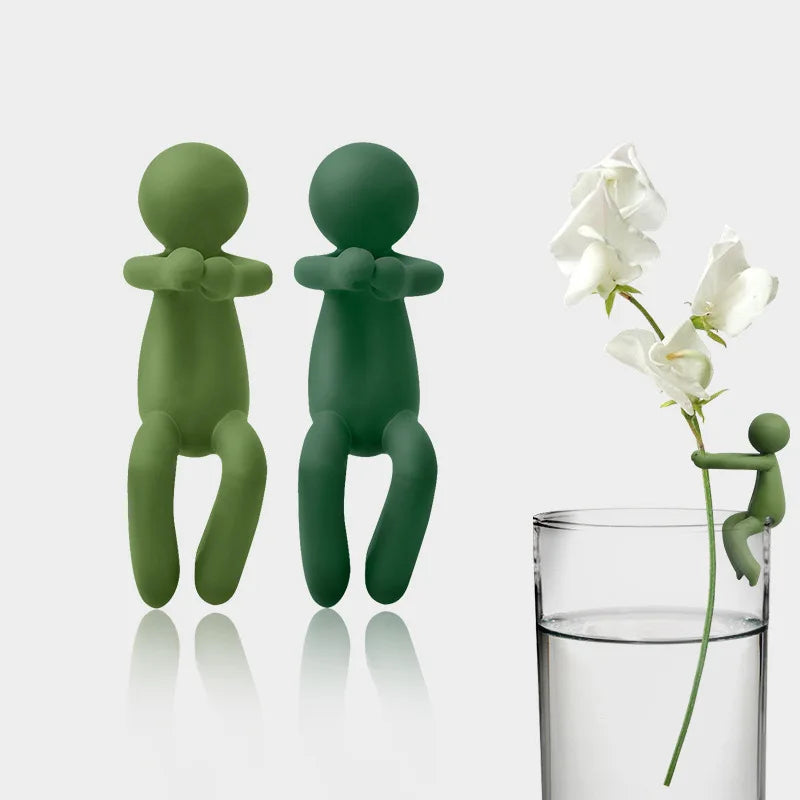 A set of three human-shaped plant supports in a muted green color standing next to a glass of water.