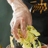 A hand wearing a clear disposable PE glove is preparing or handling green leafy vegetables in a glass bowl.