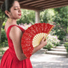 A woman in a red dress holds an open vintage lace hand fan with a gold tassel pendant outdoors.