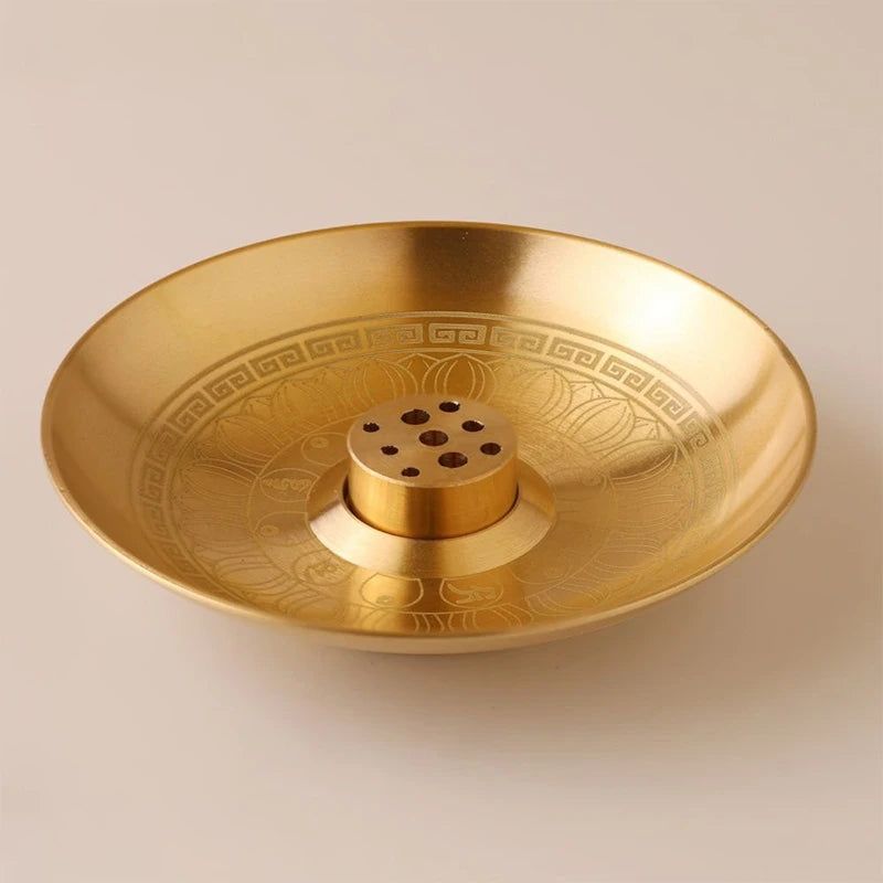 A round, brass incense stick holder plate with a decorative geometric rim and a central holder.