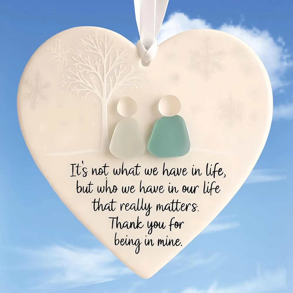 A heart-shaped sea glass ornament featuring a white tree silhouette and two small, translucent figures.