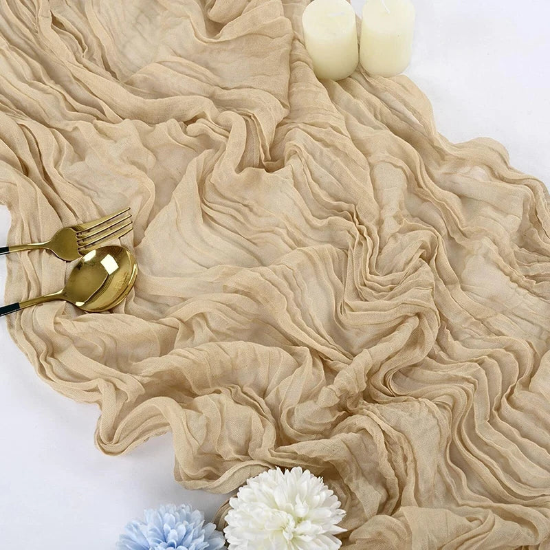 A beige gauze table runner with ruffled folds, gold cutlery, and white candles.