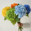 A hand holds a bouquet of silk hydrangea branches in shades of yellow, orange, and blue.