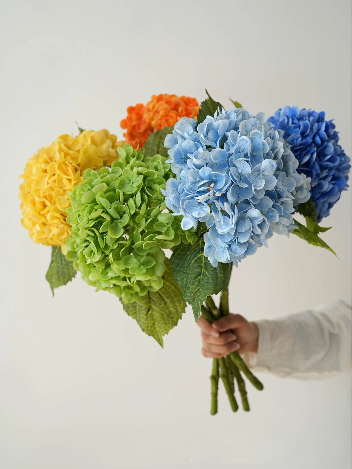 A hand holds a bouquet of silk hydrangea branches in shades of yellow, orange, and blue.