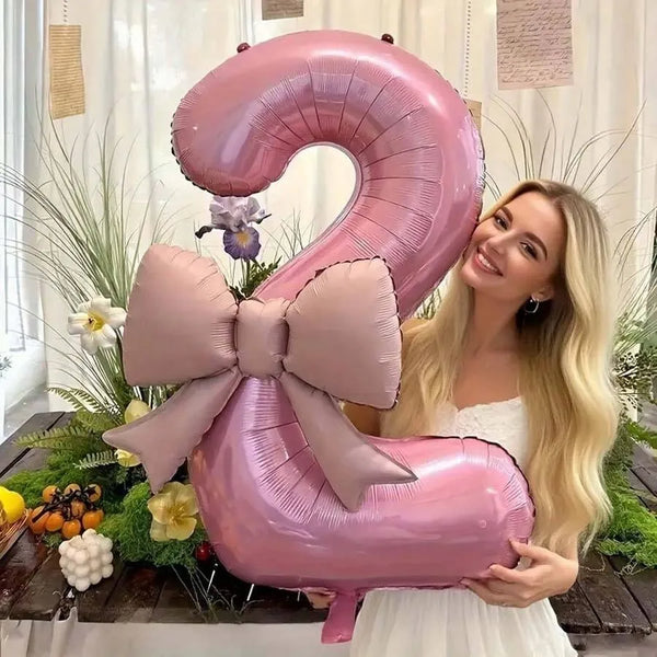 A woman holds a large pink balloon shaped like the number 2, decorated with a matte bow, in a bright indoor setting.