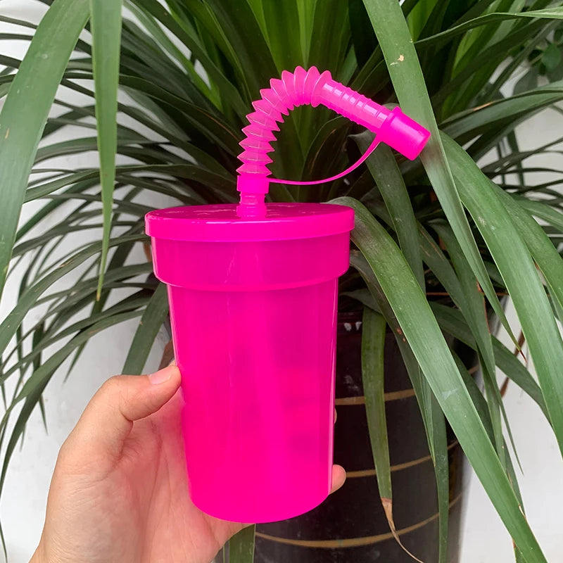 A hand holds a bright pink neon reusable sipper cup with a straw and lid against a background of green foliage.
