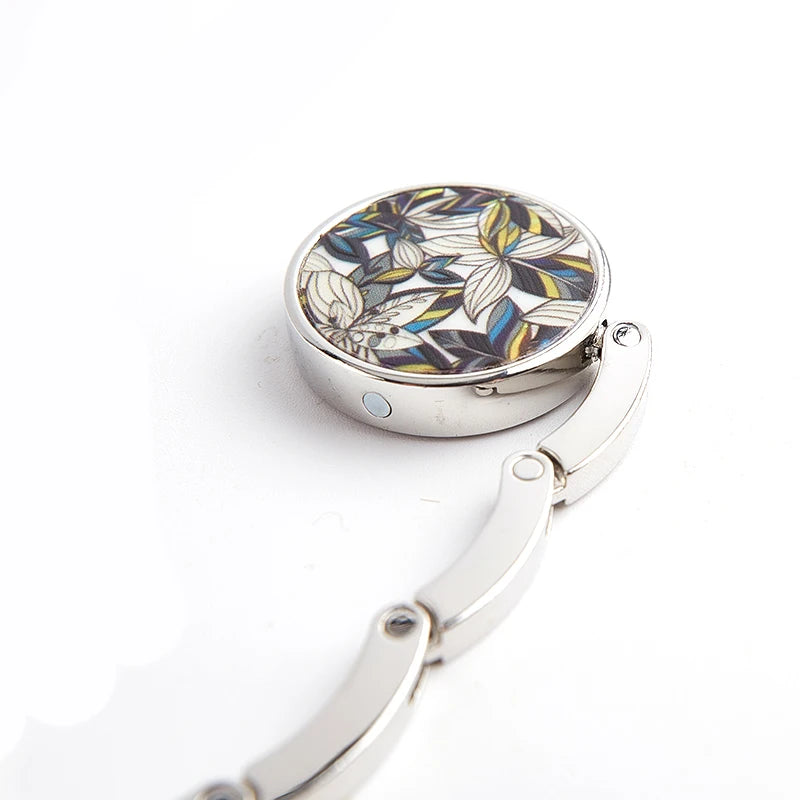 Silver foldable handbag hook with a floral patterned round plate and a curved metal handle.