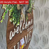 A round wooden welcome sign with white lettering and green foliage hanging against a white textured background.