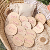 A collection of twelve wooden baby milestone cards in various shades of pink and natural wood, arranged on a beige lace fabri