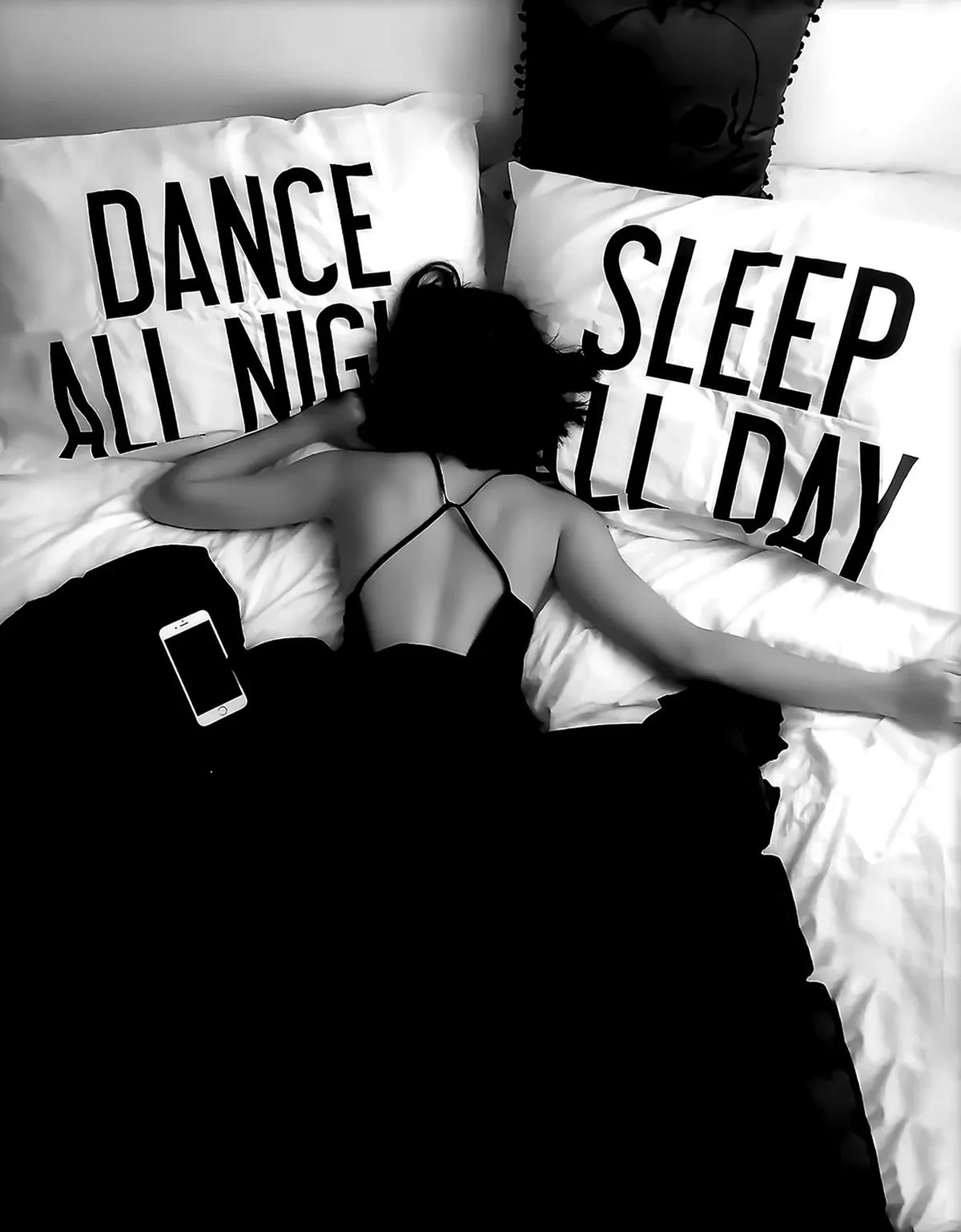 A person lying in bed with black and white text pillows reading Dance All Night and Sleep All Day.