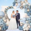 A bride and groom stand in front of a balloon arch decorated with dusty blue, sand white, and gold balloons and confetti.