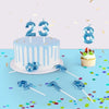 A white birthday cake decorated with blue icing and candles, surrounded by blue confetti and number toppers.