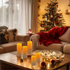 A set of four glowing yellow LED candles placed on a wooden table next to a festive Christmas tree.