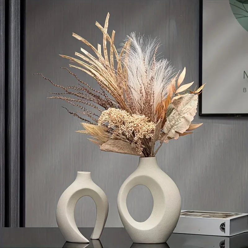 A vase arrangement featuring dried grasses and feathery plumes displayed in a pair of modern, sculptural ceramic vases.