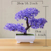 A small artificial tree bonsai decoration with purple blossoms planted in a white rectangular pot.