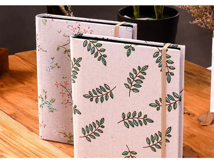 A close-up of a beige linen photo album with green botanical illustrations, resting on a wooden surface.