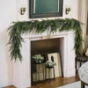 An artificial Norfolk pine garland with warm white lights draped across a white fireplace mantel.