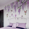 A bedroom wall decorated with watercolor wisteria tree decals featuring purple flowers, birds, and butterflies above a white 