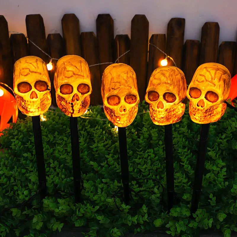 A row of glowing skull solar pathway lights decorated with green foliage and string lights.