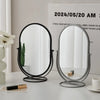 An elegant rotating oval vanity mirror with a silver and black frame sits on a white table.