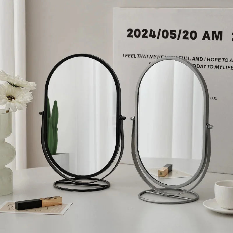 An elegant rotating oval vanity mirror with a silver and black frame sits on a white table.
