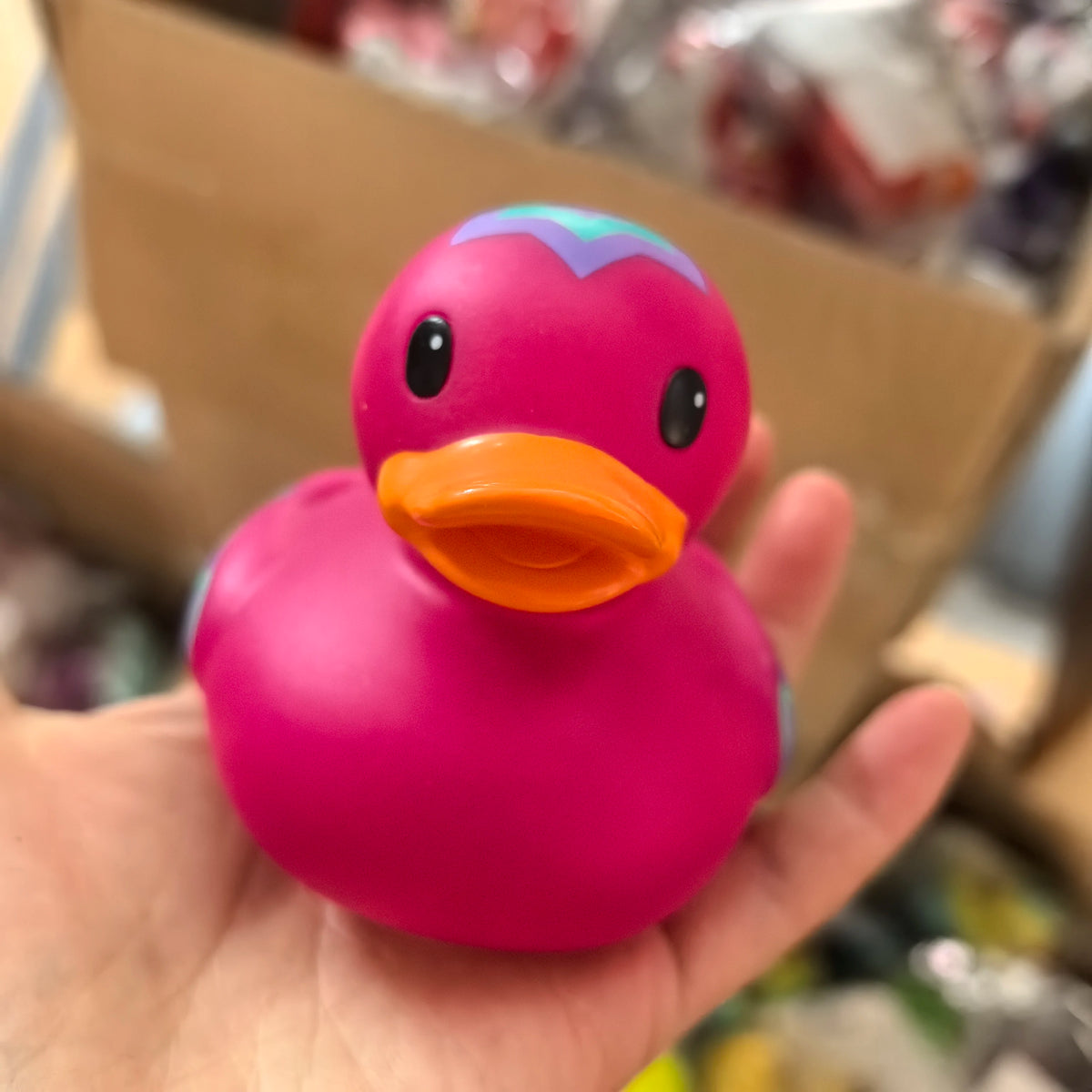 A pink rubber duck bath toy with blue accents is held in a hand against a cardboard background.