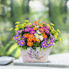 A vibrant bouquet of silk daisies in purple, orange, yellow, and white flowers displayed in a light pink pot.