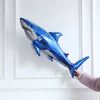 A shiny blue shark foil balloon is displayed against a white wall, held by a hand.