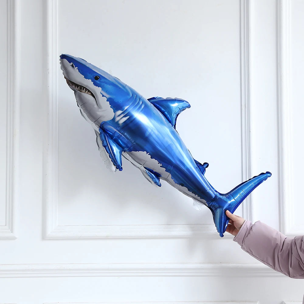 A shiny blue shark foil balloon is displayed against a white wall, held by a hand.