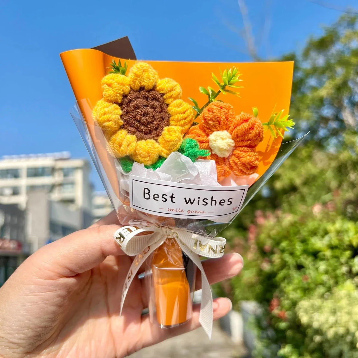 A hand holds a bouquet of handmade crochet flowers in clear cellophane with a ribbon, featuring orange and yellow blooms.