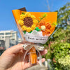 A hand holds a bouquet of handmade crochet flowers in clear cellophane with a ribbon, featuring orange and yellow blooms.