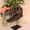 A dark resin sculpture of a realistic dinosaur skull displayed on a black stand amidst green foliage.