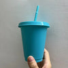 A hand holds a teal plastic cold cup with a blue lid and straw against a light gray wall.