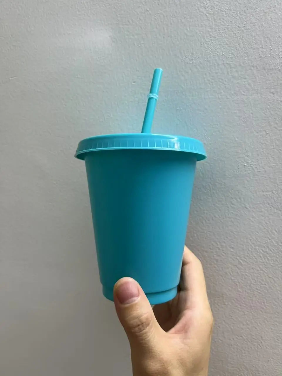 A hand holds a teal plastic cold cup with a blue lid and straw against a light gray wall.