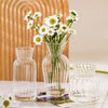 A clear glass vase filled with white daisies sits next to a tall cylindrical glass vase and a short, textured glass vase.