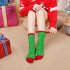 A person wearing red knitted sweater and green Christmas socks with reindeer and tree patterns sitting on a floor.