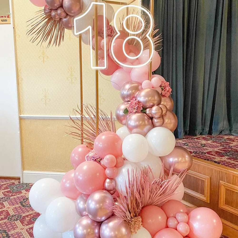 A vertical arrangement of pink and white latex balloons with metallic confetti accents against a dark curtain backdrop.